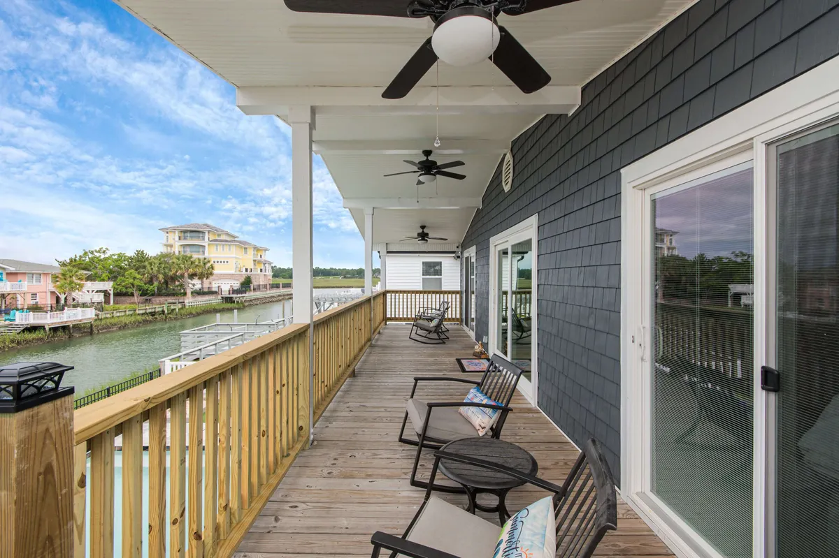 The photo showcases a spacious wooden deck overlooking the waterway, complete with comfortable seating and ceiling fans. The serene ambiance is ideal for relaxing, stargazing, or gathering for evening conversations. The surrounding houses and water views reinforce the vacation feel and outdoor living.