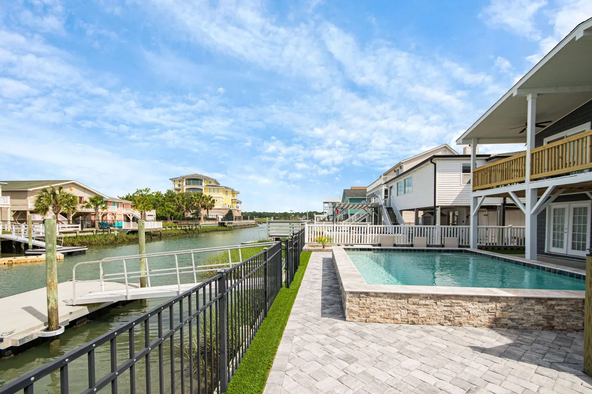An inviting exterior shot of a swimming pool beside a tranquil canal, surrounded by a patio area and nearby houses. The setting is ideal for lounging by the pool, taking a dip, or enjoying an outdoor meal while basking in the relaxing atmosphere of the waterfront.
