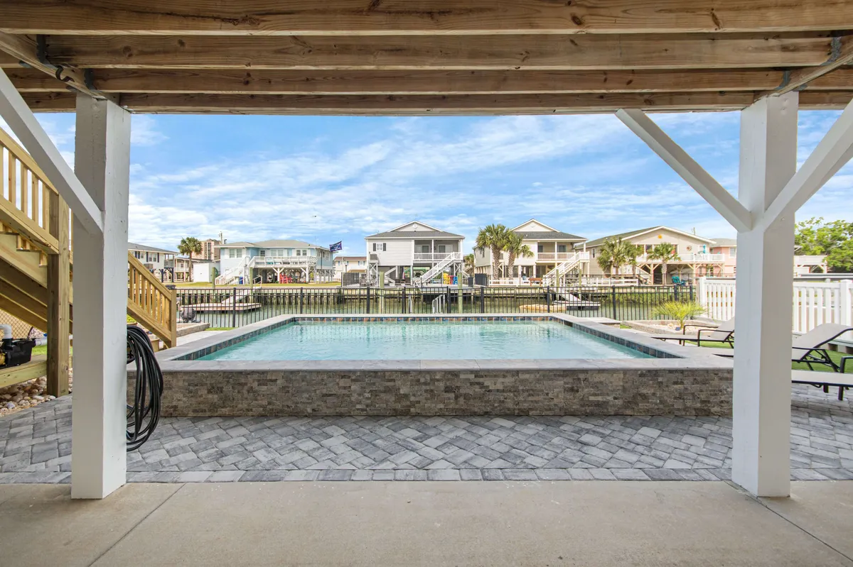 This exterior shot captures a sparkling swimming pool in the foreground with a tranquil channel just beyond, framed by lush landscaping and neighboring houses. The scene is bathed in soft, natural light, suggesting a peaceful, resort-like atmosphere ideal for both relaxation and lively gatherings. Lounge chairs and the gentle ripple of water enhance the sense of serenity and luxury.