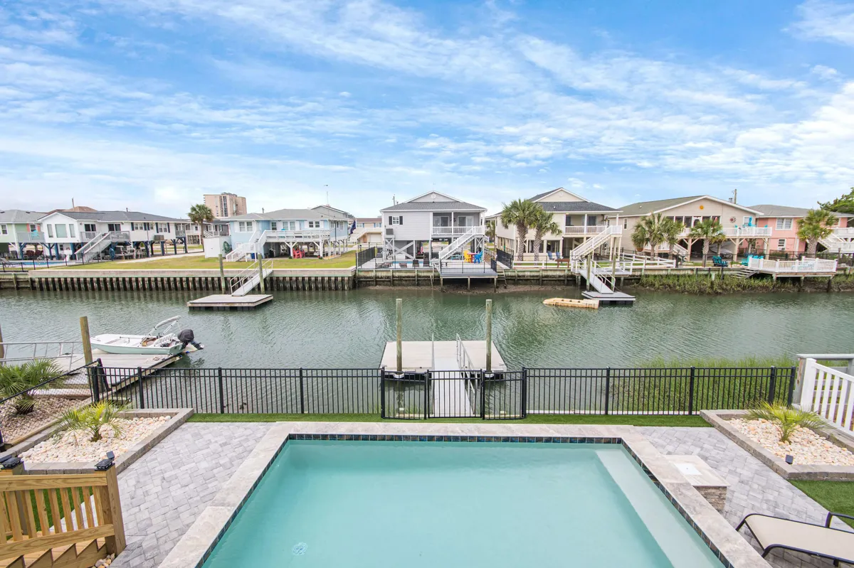 A wide exterior view featuring a pristine pool overlooking a peaceful canal, with a private dock stretching into the water. The pool area is designed for lounging and socializing, while the canal view emphasizes direct water access. Hints of outdoor dining and family-friendly spaces are visible, reinforcing the home’s versatility for both quiet mornings and lively gatherings.