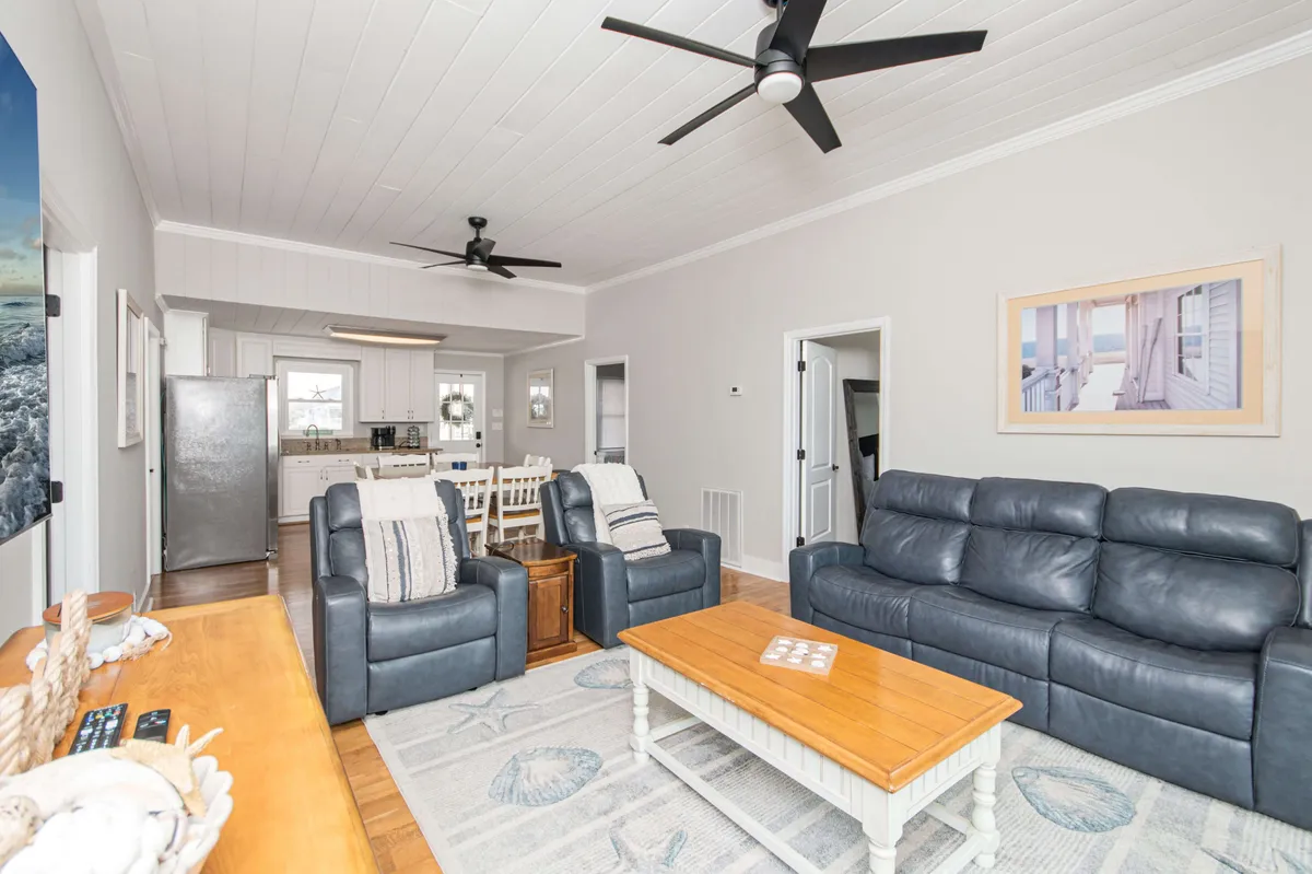 A bright, inviting living area with blue leather sofas arranged around a wooden coffee table, vibrant coastal artwork on the wall, and an open view into the adjacent gourmet kitchen area. The space is bathed in natural light, showcasing the seamless flow between living and kitchen spaces, perfect for gatherings and relaxation.