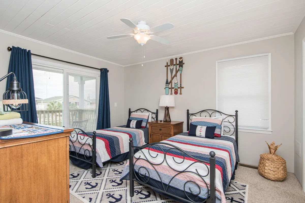 A cheerful bedroom with two twin beds featuring colorful bedding, a wooden nightstand with a lamp, a large window with curtains, and a decorative wall hanging. The room is filled with soft, natural light and an inviting, playful mood.