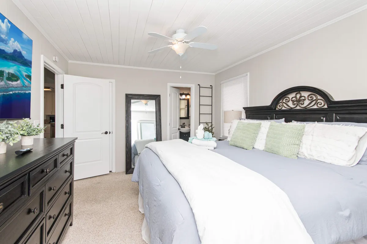 A tranquil bedroom featuring a large bed adorned with decorative pillows, a sleek black dresser, a wall mirror, and tasteful artwork. Natural light pours in from a window, illuminating the serene, uncluttered space.